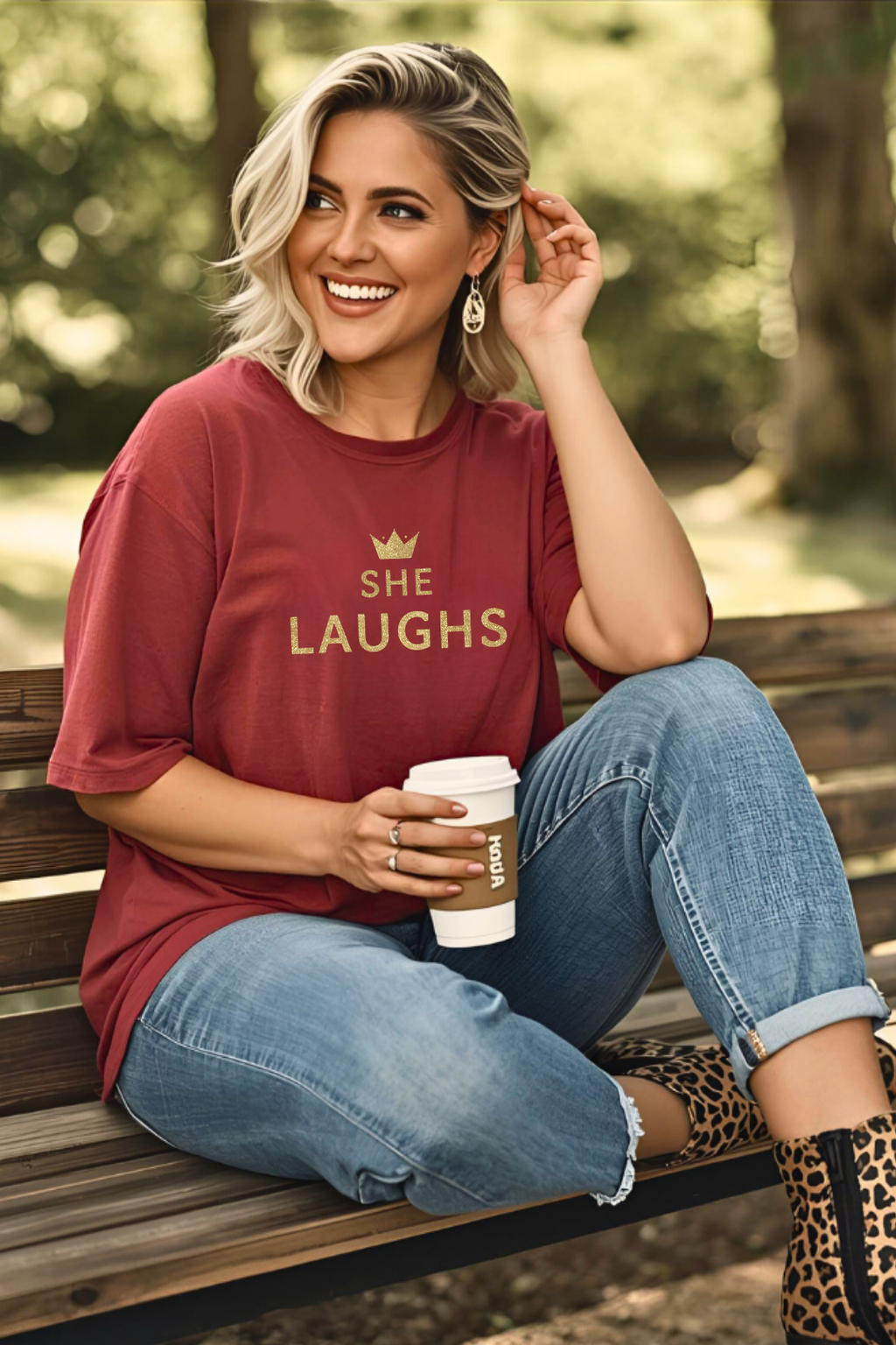 Woman wearing rust SHE LAUGHS ™ Christian tee with gold crown design while smiling on a park bench.
